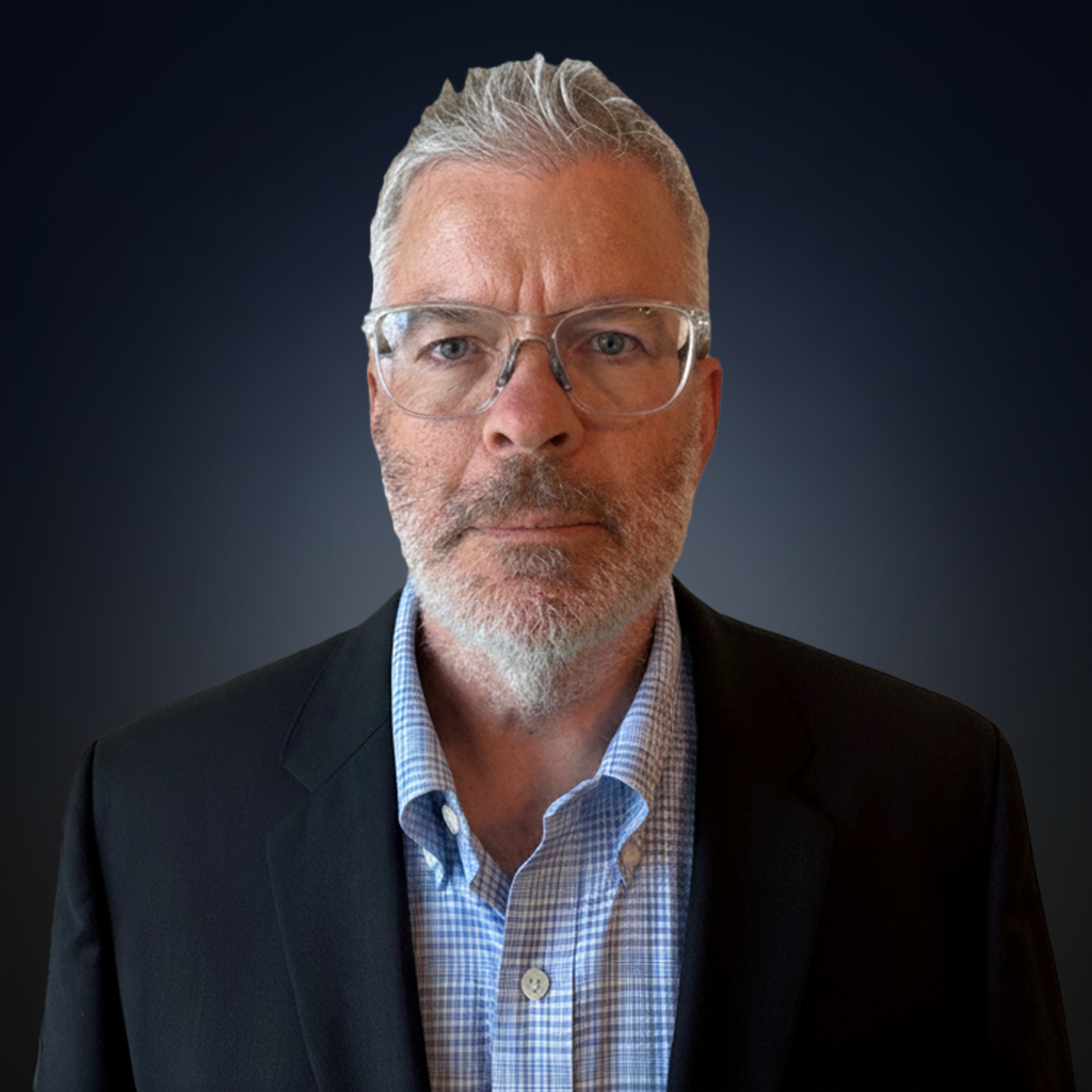 A middle-aged man with gray hair, beard, and glasses, wearing a dark suit jacket over a checked shirt, poses against a dark gradient background.