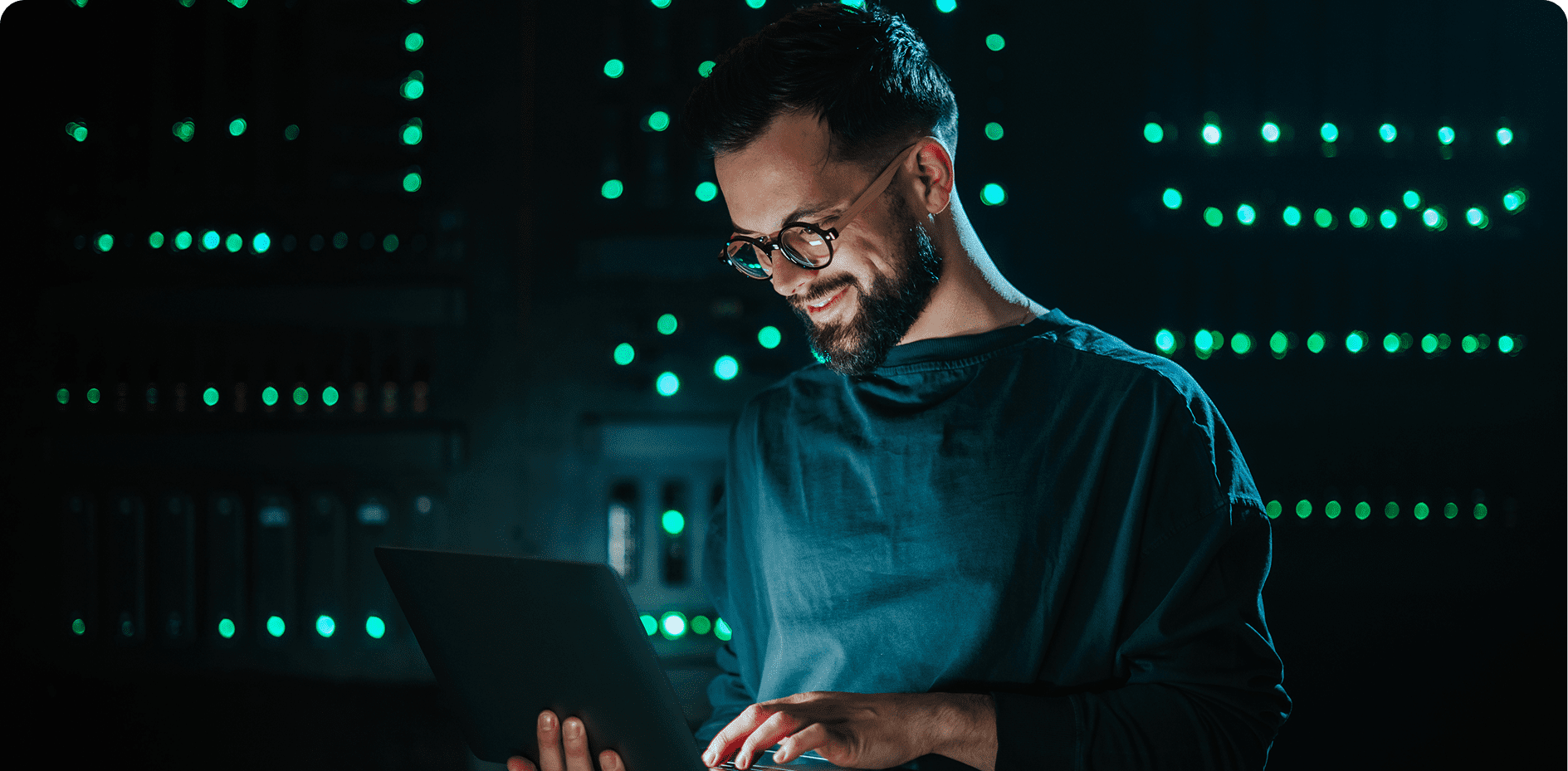 A person wearing glasses is standing in front of illuminated server racks, using a laptop and smiling.